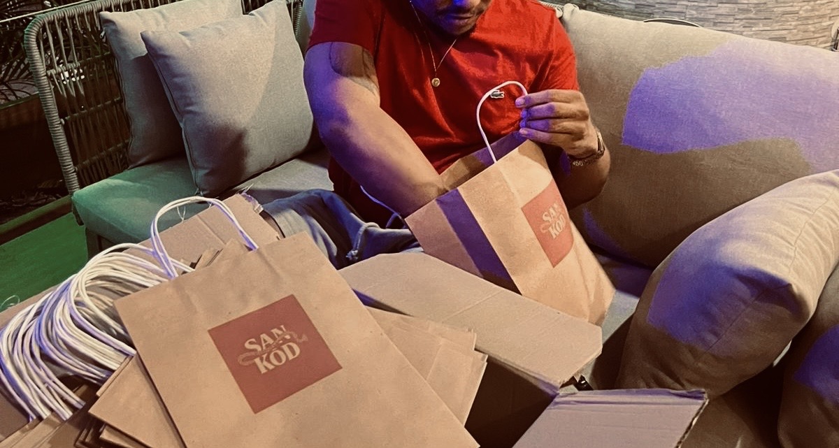 Un homme en chemise rouge prépare des sacs en papier de la marque SAN KÒD sur un canapé, entouré de boîtes en carton et de cordons.